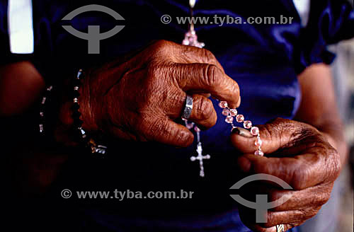  Detalhe das mãos de uma idosa rezando com um rosário com uma cruz 