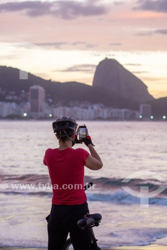 Mulher fotografando com telefone celular o nascer do sol na praia de Copacabana com o Pão de Açúcar ao fundo - Rio de Janeiro - Rio de Janeiro (RJ) - Brasil