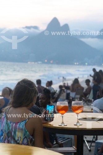 Pessoa em mesa de bar utilizando smartphone ao entardecer na praia do Arpoador com o Morro Dois Irmãos ao fundo - Rio de Janeiro - Rio de Janeiro (RJ) - Brasil