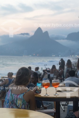 Pessoa em mesa de bar utilizando smartphone ao entardecer na praia do Arpoador com o Morro Dois Irmãos ao fundo - Rio de Janeiro - Rio de Janeiro (RJ) - Brasil