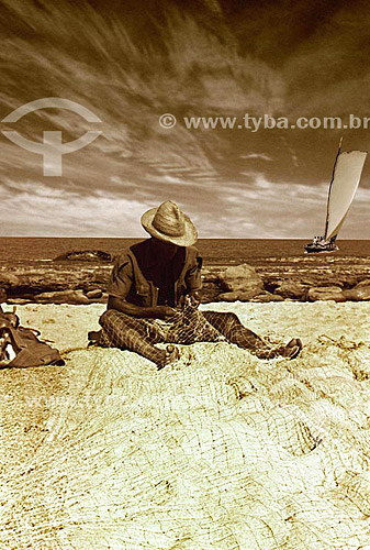  Fisherman with hat weaving fishing net with sailing boat on the sea in the background - Salvador city - Bahia state - Brazil 
