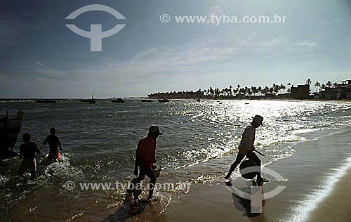  Silhouette of fishermen on the Itapoa Beach highlighted by the reflection of the sun on the water with boats and coconut trees in the background - Salvador city - Bahia state - Brazil 