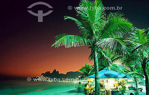  People sitting among coconut trees at a refreshment kiosk just after sunset on Ipanema Beach, with Rock of Gavea and Morro Dois Irmaos (Two Brothers Mountain) in the background - Rio de Janeiro city - Rio de Janeiro state - Brazil 