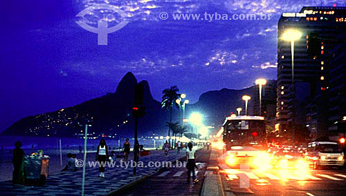  Night falls on Ipanema Beach as some people walk on the Promenade, others jog and bicycle on the bicycle path, with the lights of cars moving along Av. Vieira Souto (Vieira Souto Avenue) and the Morro Dois Irmaos (Two Brothers Mountain) in the backg 
