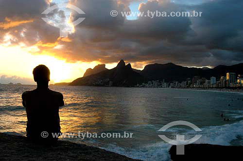  Man sitting down looking to the Ipanema Beach at sunset  - Morro Dois Irmaos (Two Brothers Mountain)* in the background - Rio de Janeiro city - Rio de Janeiro state - Brazil   *The two Brothers Mountain is National Historic Site since 08-08-1973. 