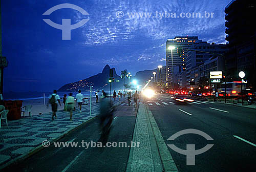  View of Ipanema Beach at sunset with Morro Dois Irmãos (Two Brothers Mountain) in the background, people walking on the sidewalk and bicycling and jogging on the bicycle path, and the lights of cars passing along Av. Vieira Souto (Vieira Souto Avenu 