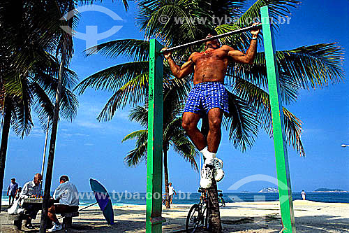  Man exercising on a chin-up bar on Ipanema Beach with two older men playing chess to the left - Rio de Janeiro state - Brazil 