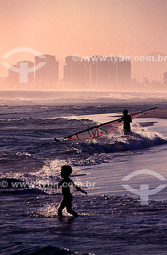  Silhouette of a child and a windsurfer in the water on Barra da Tijuca Beach at sunset, with buildings in the background - Rio de Janeiro city - Rio de Janeiro state - Brazil 