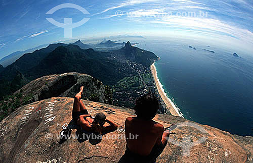  A young couple sitting atop Rock of Gavea* admiring the view of Sao Conrado Beach below to the right, at the foot of Morro Dois Irmaos (Two Brothers Mountain) - Rio de Janeiro city - Rio de Janeiro state - Brazil  *The Rock of Gavea is a National Hi 