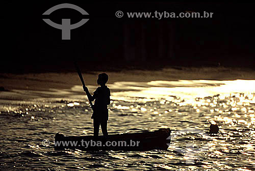  Silhouette of a boy rowing a boat made out of a tree trunk, highlighted by the reflections of the setting sun on the water - Angra dos Reis city - Costa Verde (Green Coast) - Rio de Janeiro state - Brazil 