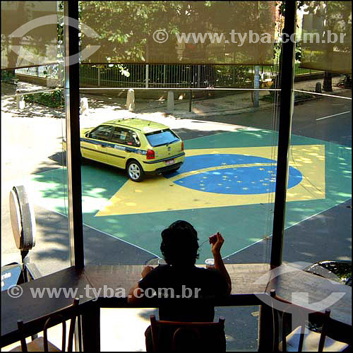  Woman eating in a restaurant in the foreground with a taxi passing over a Brazil flag painted on the road - Ipanema  neighborhood - Rio de Janeiro city - Rio de Janeiro state - Brazil 