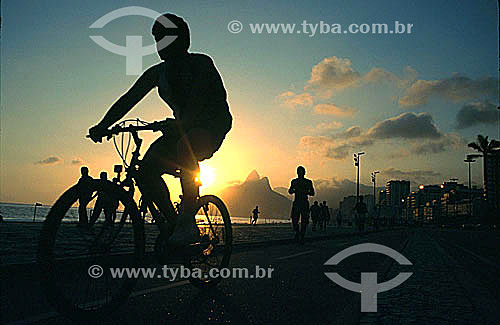  Silhueta de ciclista na ciclovia da Praia de Ipanema e pessoas caminhando e correndo ao pôr-do-sol com o Morro Dois Irmãos ao fundo - Rio de Janeiro RJ - Brasil  - Rio de Janeiro - Rio de Janeiro - Brasil