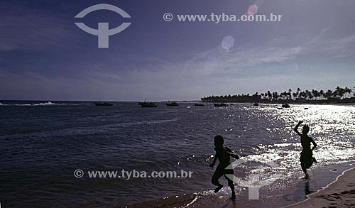  Silhouette of two boys running on the Itapoa Beach highlighted by the reflection of the sun on the water with coconut trees in the background - Salvador city - Bahia state - Brazil 